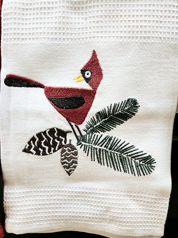 A dish towel featuring a Northern Cardinal bird on a pine branch with pine cones.